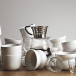 Chaos blank white and transparent coffee cups on thick wooden table. Drip coffee maker with filtered beverage in center.